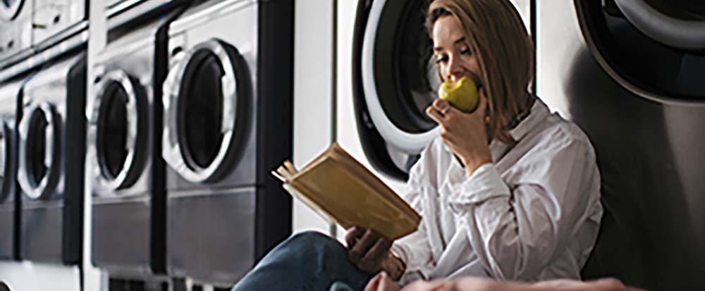 A woman eating an apple while sitting on the floor in front of a row of laundry machines