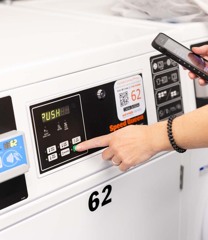 A hand reaches to press a button on a laundry machine while the other hand uses a phone to access the LaundryConnect app