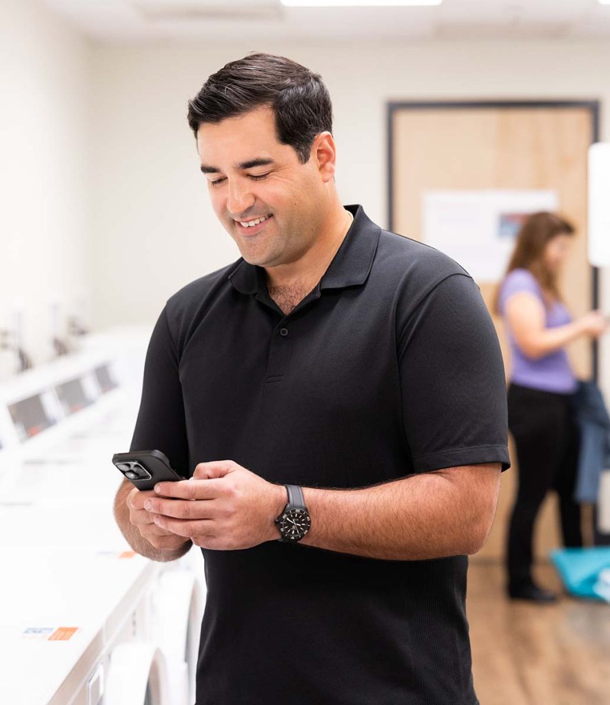 A man stands in a laundry room smiling while using his phone to access the WashWallet app