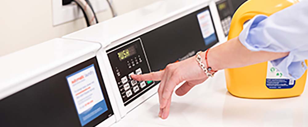 A hand reaches for a button on a laundry machine with detergent sitting next to it