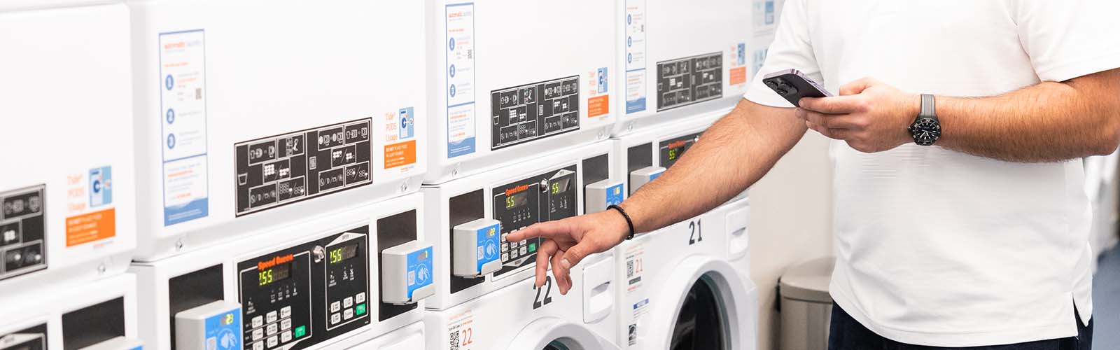 A hand reaches to press a button on a laundry machine while the other hand uses a phone to access the laundry resources app, LaundryConnect